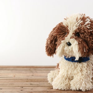 May include: A brown and white yarn dog toy with a blue collar. The dog is sitting on a wooden surface.
