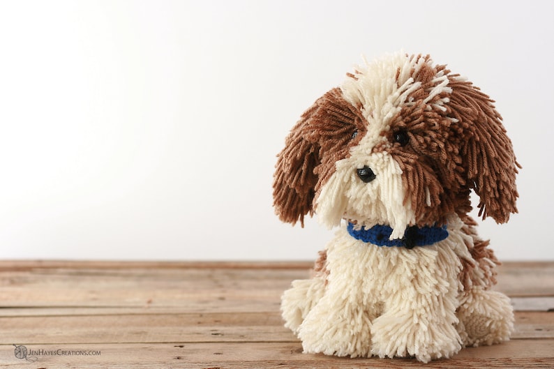 May include: A brown and white yarn dog toy with a blue collar. The dog is sitting on a wooden surface.