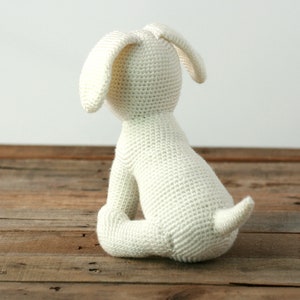 May include: A white crocheted stuffed dog sitting on a wooden surface. The dog is facing away from the camera, with its tail curled up behind it.
