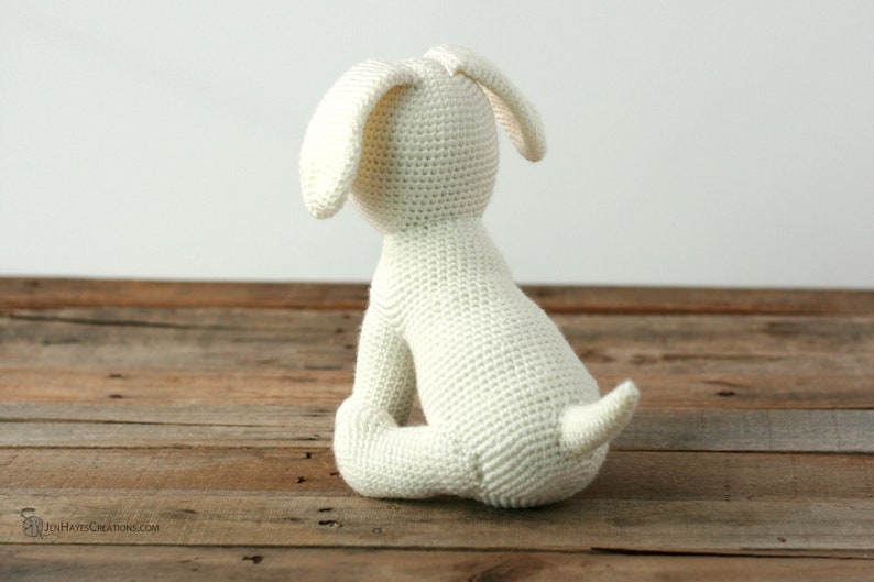 May include: A white crocheted stuffed dog sitting on a wooden surface. The dog is facing away from the camera, with its tail curled up behind it.