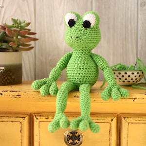 May include: A green crocheted frog with white eyes sits on a yellow dresser with two drawers. The frog has long legs and a wide smile.