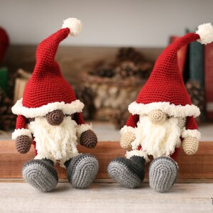 A brown and a white Crochet Santa Gnomes sit side by side on a long piece of wood. Pine cones, yarn, books and small packages are blurred in the background.