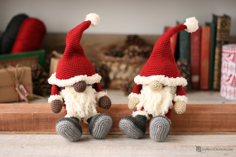 A brown and a white Crochet Santa Gnomes sit side by side on a long piece of wood. Pine cones, yarn, books and small packages are blurred in the background.