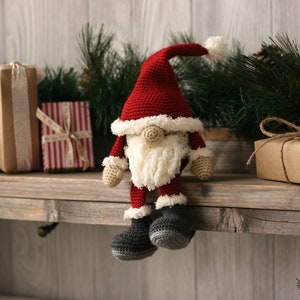 A Crochet Santa Gnome sitting on a wooden mantel surrounded by pine garland and rustic packages.