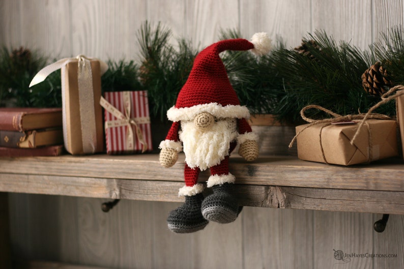 A Crochet Santa Gnome sitting on a wooden mantel surrounded by pine garland and rustic packages.