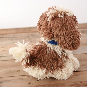 May include: A brown and white yarn dog toy with a blue collar. The toy is sitting on a wooden surface.