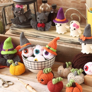 Op de afbeelding: Een verzameling gehaakte Halloween-thema amigurumi-speelgoed, waaronder pompoenen, geesten, vleermuizen en oogballen. Het speelgoed is gerangschikt op een houten tafel met garen, een kom en andere knutselbenodigdheden.