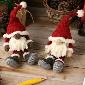 A brown and a white Crochet Santa Gnome sitting side by side surrounded by pinecones, packages, and other Christmas paraphernalia.