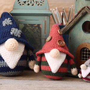 May include: Three crocheted gnomes, one in blue and gray stripes, one in red and green stripes, and one in red and gray stripes. The gnomes are sitting on a wooden surface in front of a wooden birdhouse.
