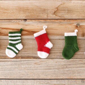 May include: Five miniature crocheted Christmas stockings in green, white, and red. The stockings are hanging on a wooden surface.