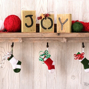 Three Mini Crochet Stockings in red, white, and green variations hanging from a mantle with decorations that say JOY and skeins and balls of green, red, and white yarn.