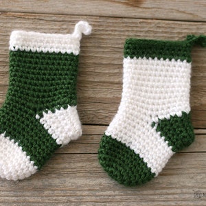 Two Mini Crocheted Stockings in green with a white heel, toe, and top band and white with a green heel, toe, and top band lying on a wooden background.