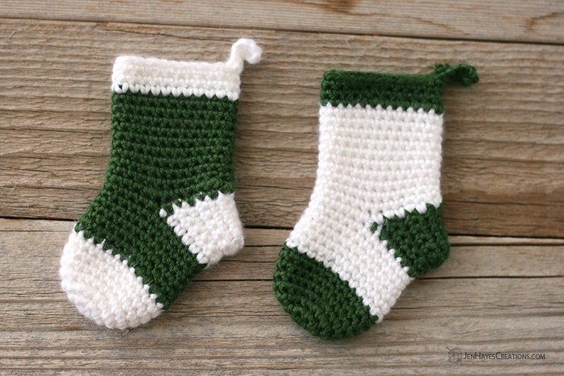Two Mini Crocheted Stockings in green with a white heel, toe, and top band and white with a green heel, toe, and top band lying on a wooden background.
