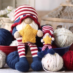 Può includere: Due gnomi all'uncinetto con colori patriottici. Lo gnomo più grande tiene una stella gialla e indossa un cappello rosso, bianco e blu e gambe a righe. Lo gnomo più piccolo tiene anche una stella. Gomitoli di lana rossi, bianchi e blu sullo sfondo.