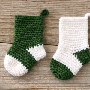 Two Mini Crocheted Stockings in green with a white heel and toe and white with a green heel and toe lying on a wooden background.