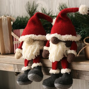 Side angle view of a brown and a white Crochet Santa Gnome sitting beside each other on a wooden mantel surrounded by pine garland and rustic packages.