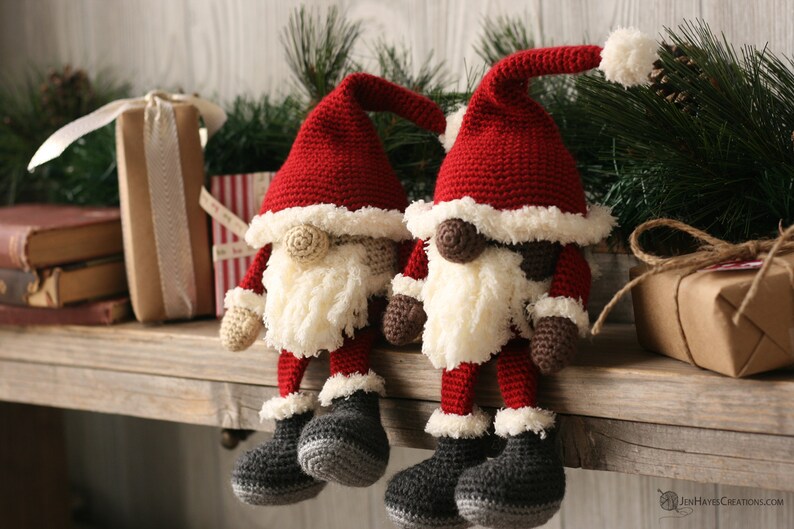 Side angle view of a brown and a white Crochet Santa Gnome sitting beside each other on a wooden mantel surrounded by pine garland and rustic packages.