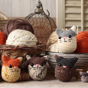 May include: Five crocheted cat toys in various colors, including gray, brown, and orange. The toys are sitting on a table with yarn balls and other crafting supplies in the background.