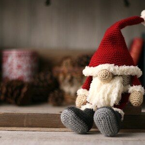 Crochet Santa Gnome sitting on the right side of a long piece of wood. Pine cones, yarn, books and small packages blurred in the background.