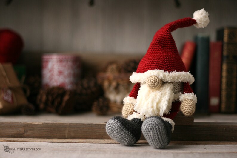 Crochet Santa Gnome sitting on the right side of a long piece of wood. Pine cones, yarn, books and small packages blurred in the background.