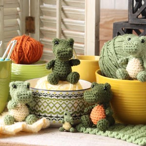 Puede incluir: Una colección de juguetes de cocodrilo hechos a mano con ganchillo en varios tamaños y poses. Los cocodrilos son principalmente verdes con vientres color crema y acentos naranjas. Se exhiben con hilo, ganchos de ganchillo y cuencos decorativos.