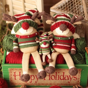 May include: Three crocheted reindeer ornaments in a green wooden box with the text "Happy Holidays". The reindeer are wearing red and green striped hats and sweaters. The box is filled with yarn and crochet hooks.