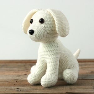 May include: A white crocheted stuffed dog sitting on a wooden surface. The dog has black eyes and a black nose.