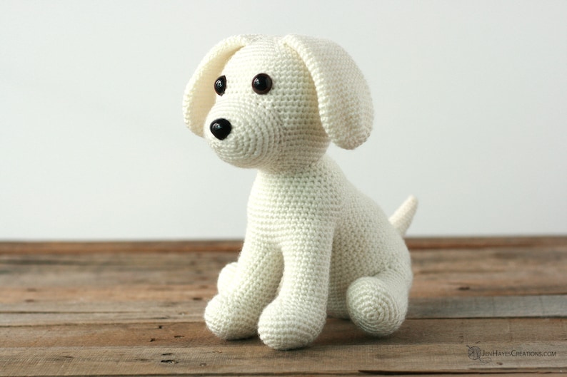 May include: A white crocheted stuffed dog sitting on a wooden surface. The dog has black eyes and a black nose.