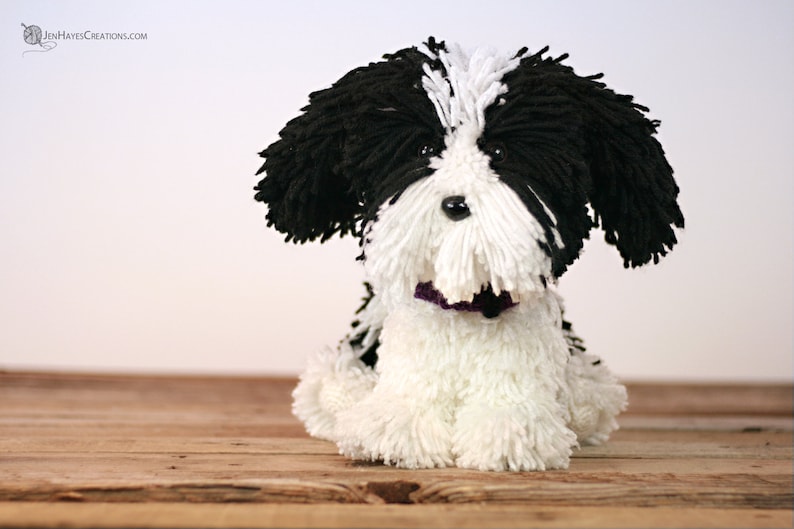 May include: A black and white yarn dog toy with a purple collar. The dog is sitting on a wooden surface.