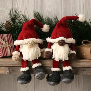 A white and a brown Crochet Santa Gnome sitting beside each other on a wooden mantel surrounded by pine garland and rustic packages.