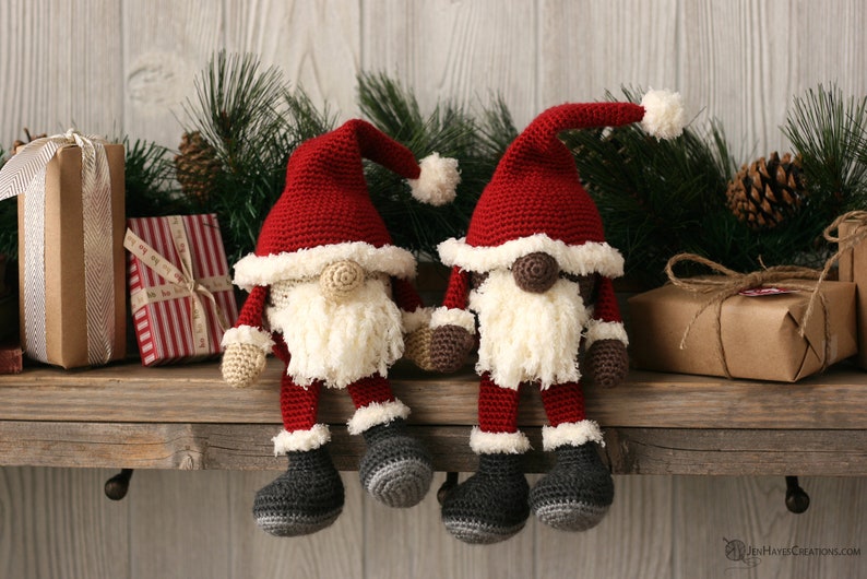 A white and a brown Crochet Santa Gnome sitting beside each other on a wooden mantel surrounded by pine garland and rustic packages.