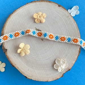 May include: A beaded bracelet with a repeating pattern of orange and blue flower-like shapes on a white background. The bracelet is laid on a wooden slice with decorative paper flowers on a blue surface. The bracelet has a silver clasp.