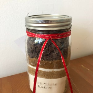 以下が含まれることがあります： チョコレートチップなど、クッキーを焼くための材料が層状に入った透明なガラスのメイソンジャー。ジャーは銀色の蓋で密閉され、赤いリボンで結ばれています。ジャーは木製の表面に置かれています。