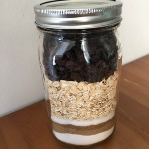 以下が含まれることがあります： 銀色の蓋が付いた透明なガラス瓶で、焼き菓子の材料が層状に詰められています。層には、濃いレーズン、オートミール、白砂糖、ブラウンシュガー、さらに白砂糖が含まれています。瓶は木製の表面に置かれています。