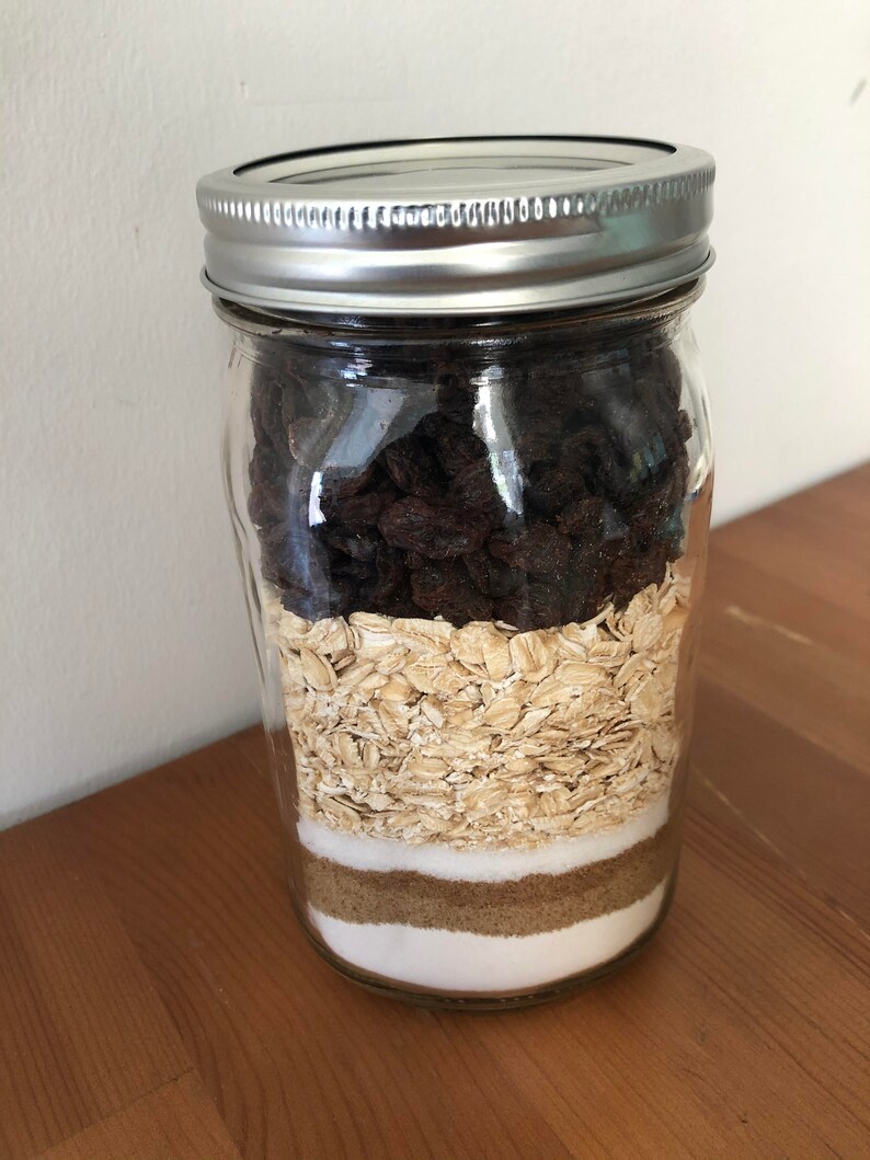 以下が含まれることがあります： 銀色の蓋が付いた透明なガラス瓶で、焼き菓子の材料が層状に詰められています。層には、濃いレーズン、オートミール、白砂糖、ブラウンシュガー、さらに白砂糖が含まれています。瓶は木製の表面に置かれています。
