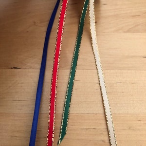 以下が含まれることがあります： さまざまな色の装飾用リボンのスプール。リボンには、青、金色のトリム付きの赤、金色のトリム付きの緑、金色のトリム付きのクリーム色が含まれます。リボンは、明るい茶色の木製の表面に表示されています。