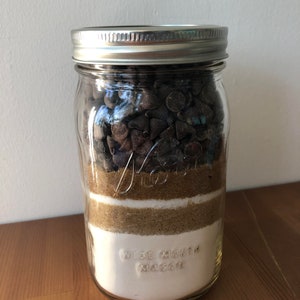 以下が含まれることがあります： チョコレートチップクッキーの材料が層状に入った透明なガラスのメイソンジャー。層には、チョコレートチップ、ブラウンシュガー、白砂糖、小麦粉が含まれています。ジャーには銀色の蓋があり、「WIDE MOUTH MASON」の文字がエンボス加工されています。