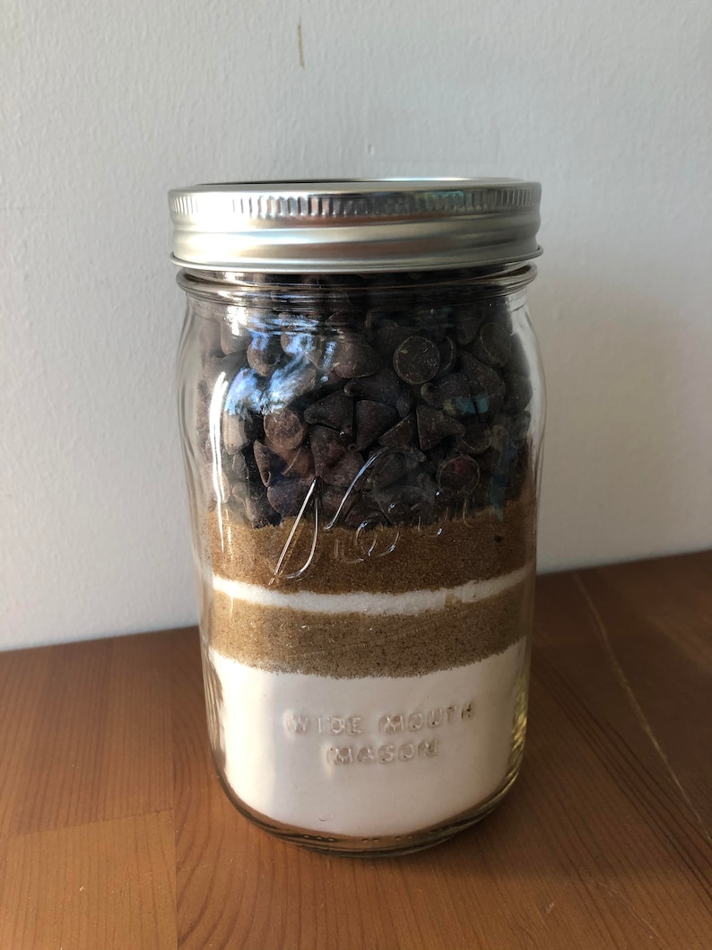 以下が含まれることがあります： チョコレートチップクッキーの材料が層状に入った透明なガラスのメイソンジャー。層には、チョコレートチップ、ブラウンシュガー、白砂糖、小麦粉が含まれています。ジャーには銀色の蓋があり、「WIDE MOUTH MASON」の文字がエンボス加工されています。