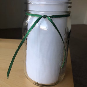 以下が含まれることがあります： 中央に緑のリボンを結んだ透明なガラス瓶。リボンは金色のトリムが付いています。瓶は白い紙で満たされています。瓶は明るい茶色の木製の表面に置かれています。