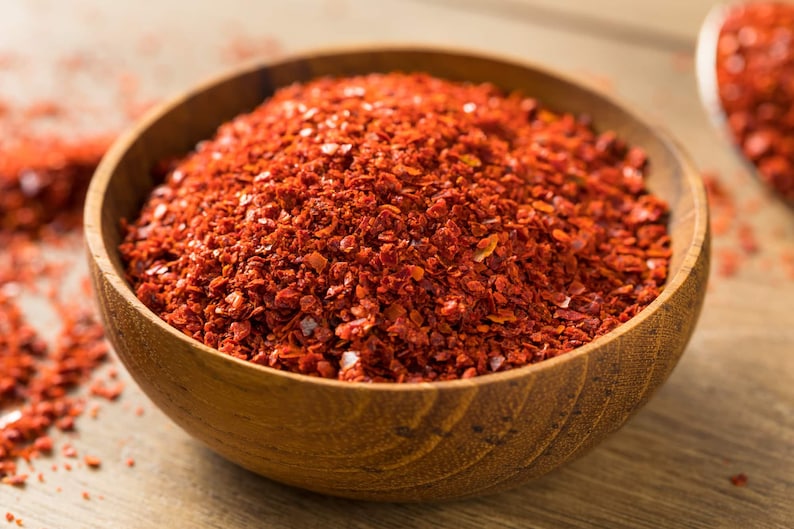 May include: A wooden bowl filled with coarsely ground, bright red chili flakes. The flakes are piled high in the bowl, with some spilling onto the wooden surface. The bowl is made of light brown wood.