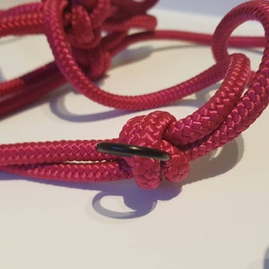 Puede incluir: Primer plano de una cuerda rosa con un anillo de metal negro. La cuerda está atada en un nudo y el anillo está unido al nudo.