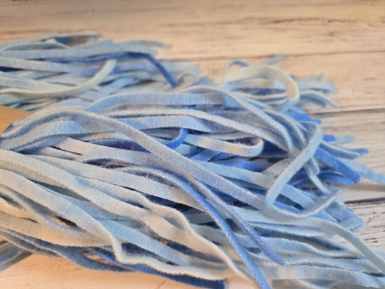 May include: A collection of thin, flexible strips in various shades of blue, ranging from pale sky blue to deep royal blue. The strips have a soft, felt-like texture and are arranged in a dense, overlapping cluster, creating a layered appearance.