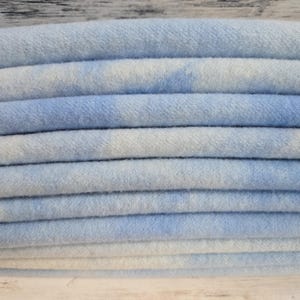 May include: A stack of folded fabric featuring a soft, light blue and white tie-dye pattern. The material has a visible, slightly fuzzy texture, and the layers are neatly arranged to show the repeating cloud-like colour variations across the surface.