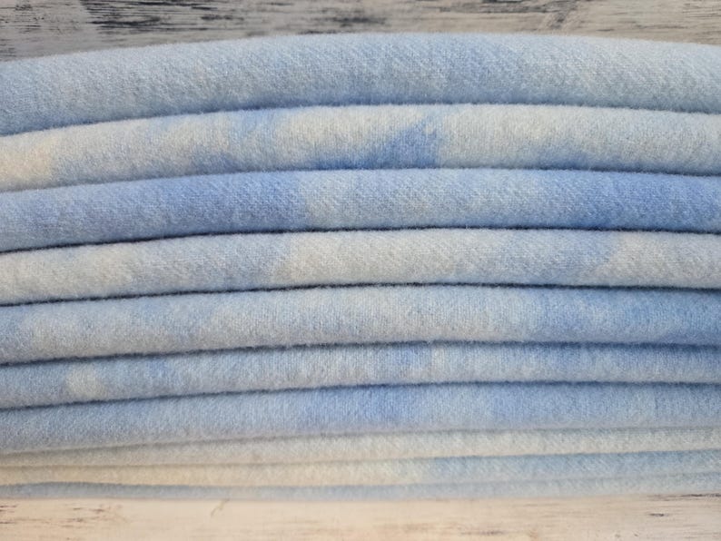 May include: A stack of folded fabric featuring a soft, light blue and white tie-dye pattern. The material has a visible, slightly fuzzy texture, and the layers are neatly arranged to show the repeating cloud-like colour variations across the surface.