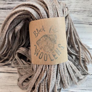 May include: A bundle of gray wool strips wrapped with a brown paper label. The label reads "Black Leg Woolens" with a dog silhouette. The wool strips are a neutral color, suitable for various craft projects.