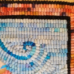 May include: A hand-hooked rug fragment featuring a textured surface of looped wool yarn. The design includes a central spiral pattern in shades of light blue and bright blue, bordered by a thick black line and a multi-coloured edge of pink, orange, and red.
