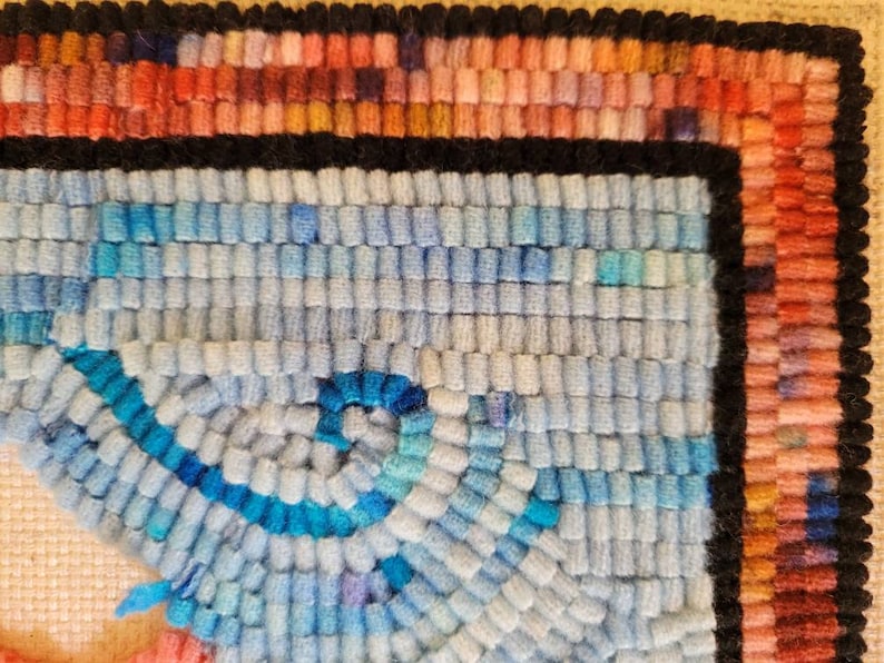 May include: A hand-hooked rug fragment featuring a textured surface of looped wool yarn. The design includes a central spiral pattern in shades of light blue and bright blue, bordered by a thick black line and a multi-coloured edge of pink, orange, and red.