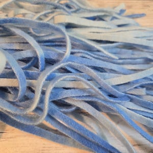 May include: A collection of thin, flexible strips of velvet ribbon in various shades of blue, ranging from pale sky blue to deep azure. The strips are arranged in a loose, overlapping pile, showcasing a soft, fuzzy texture and a consistent width.