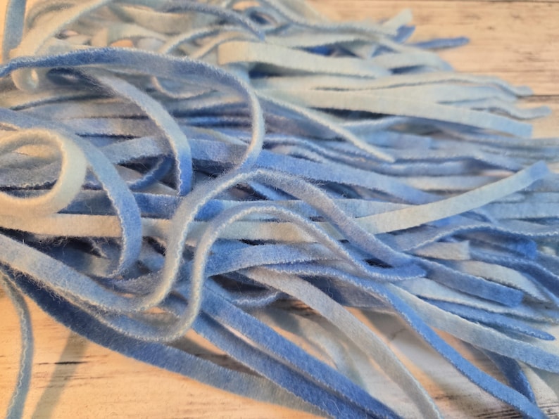 May include: A collection of thin, flexible strips of velvet ribbon in various shades of blue, ranging from pale sky blue to deep azure. The strips are arranged in a loose, overlapping pile, showcasing a soft, fuzzy texture and a consistent width.
