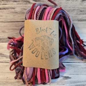 May include: A bundle of colorful wool strips in shades of pink, red, purple, and brown. The wool is wrapped around a cardboard tube with the text "Black Lab Woolens" and a dog illustration.